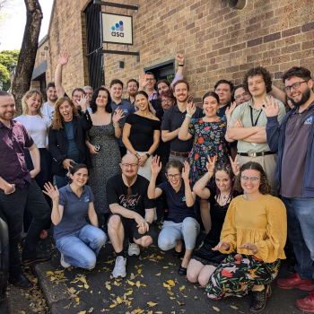 A large group of people posing outside a brick building, smiling, waving, and celebrating in a team gathering.